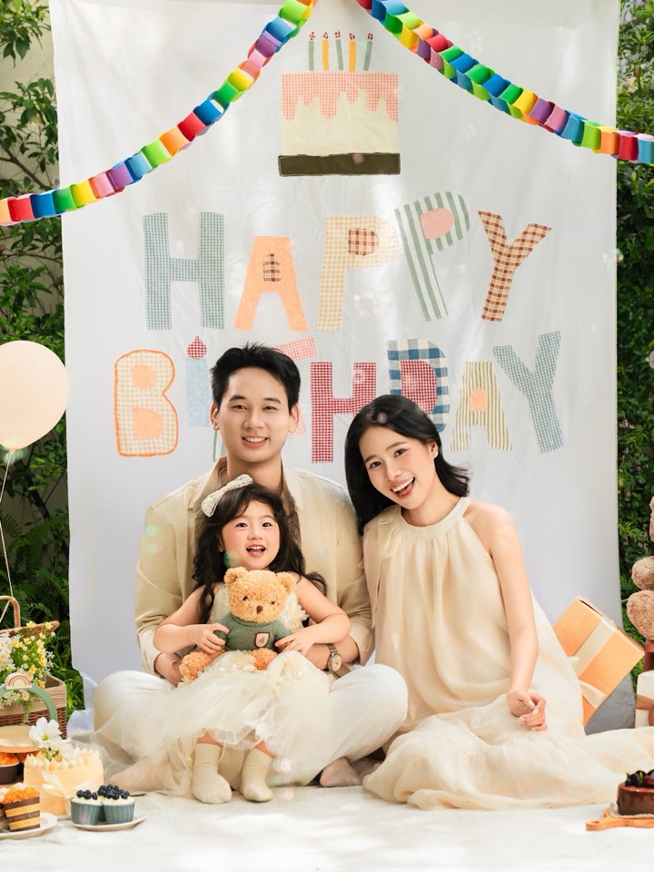 Ba mẹ và bé gái ngồi cùng nhau trước phông Happy Birthday, có bóng bay và gấu bông — concept Gia đình tại Aloha.