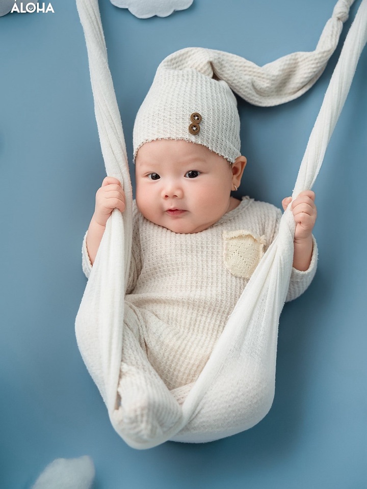 Bé sơ sinh mặc đồ len kem, đội mũ chóp, nằm trong võng vải trên nền xanh nhạt — concept Newborn tại Aloha.